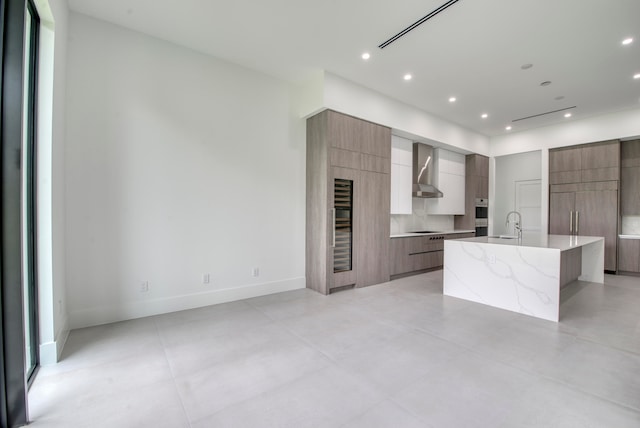 kitchen with light stone counters, wall chimney exhaust hood, an island with sink, electric stovetop, and tasteful backsplash