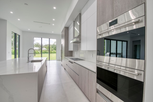 kitchen with light stone countertops, sink, wall chimney exhaust hood, double oven, and a kitchen island with sink