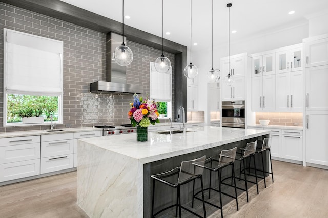 kitchen with a kitchen island with sink, wall chimney range hood, sink, and white cabinets