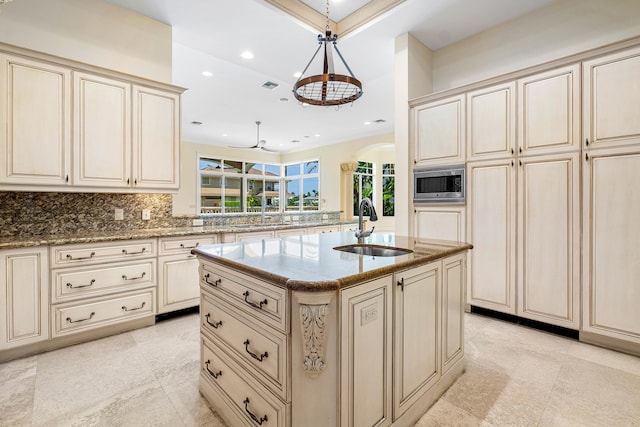 kitchen featuring sink, cream cabinets, decorative light fixtures, stainless steel microwave, and an island with sink