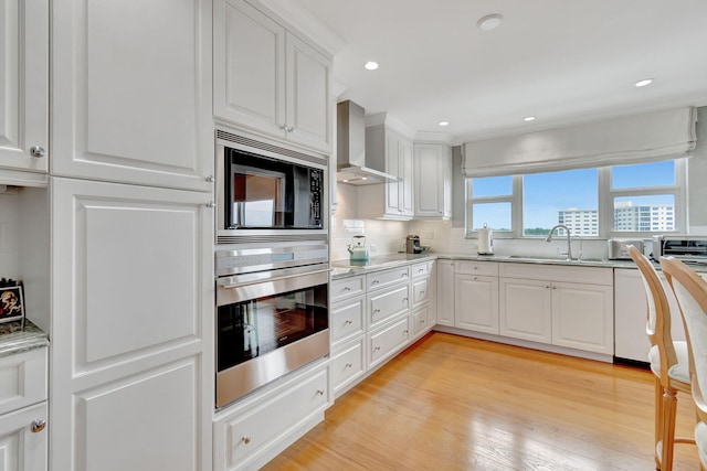 kitchen with white cabinetry, sink, wall chimney range hood, light hardwood / wood-style flooring, and appliances with stainless steel finishes