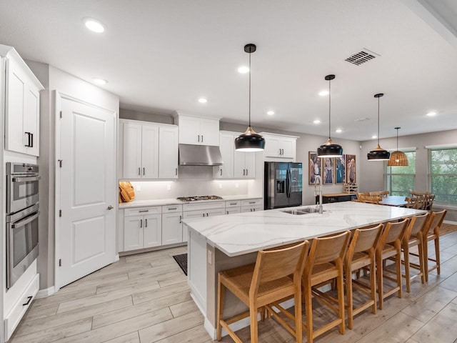 kitchen with a large island with sink, stainless steel appliances, hanging light fixtures, white cabinets, and light hardwood / wood-style floors