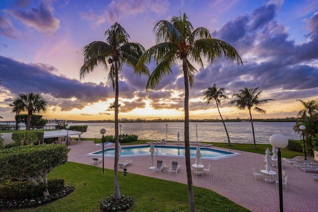 pool at dusk featuring a lawn, a water view, and a patio