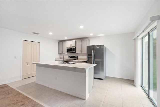 kitchen with a textured ceiling, sink, an island with sink, appliances with stainless steel finishes, and gray cabinetry