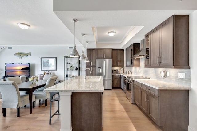 kitchen with sink, light hardwood / wood-style floors, stainless steel appliances, wall chimney exhaust hood, and a kitchen island with sink