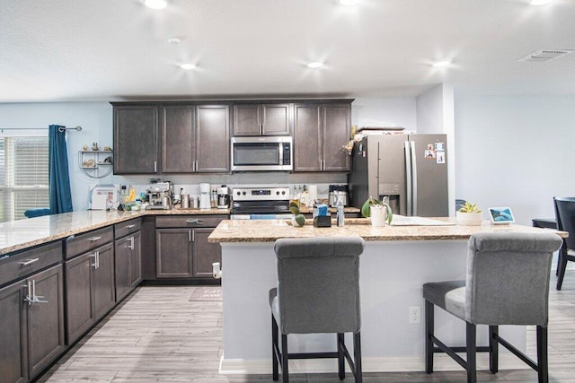 kitchen with dark brown cabinets, light hardwood / wood-style flooring, appliances with stainless steel finishes, a center island, and a kitchen bar