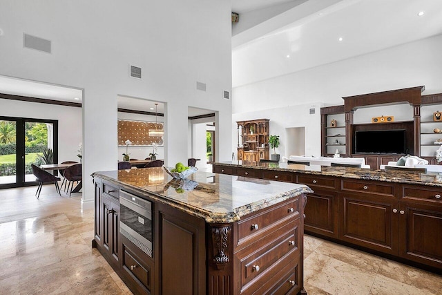 kitchen with a towering ceiling, stainless steel microwave, dark brown cabinets, a center island, and dark stone counters