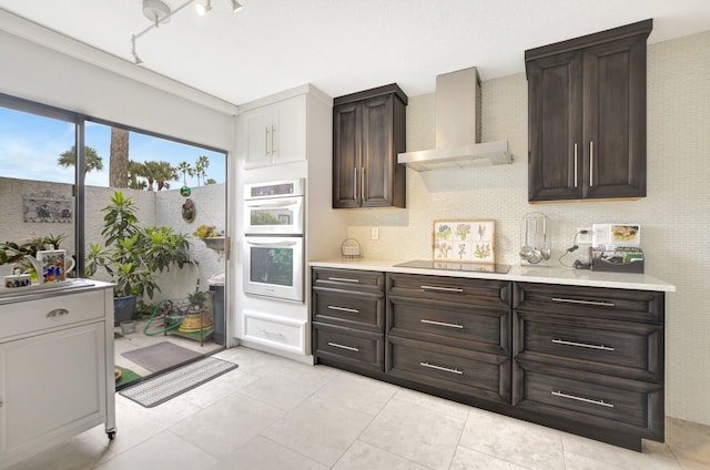 kitchen with stainless steel double oven, black electric cooktop, dark brown cabinetry, wall chimney range hood, and light tile patterned flooring