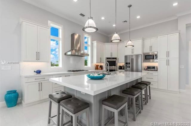 kitchen with wall chimney range hood, white cabinets, appliances with stainless steel finishes, and an island with sink