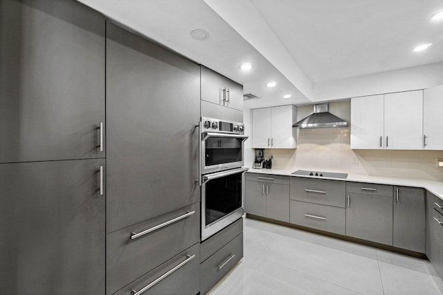 kitchen with black electric cooktop, decorative backsplash, white cabinets, wall chimney exhaust hood, and stainless steel double oven