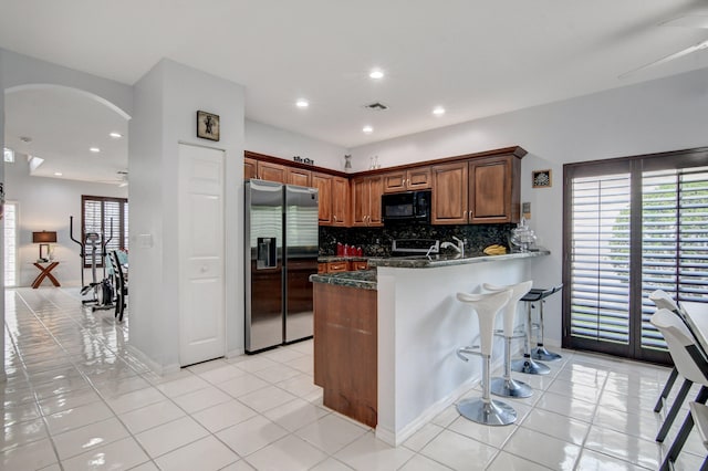 kitchen featuring kitchen peninsula, backsplash, dark stone counters, stainless steel fridge with ice dispenser, and light tile patterned floors