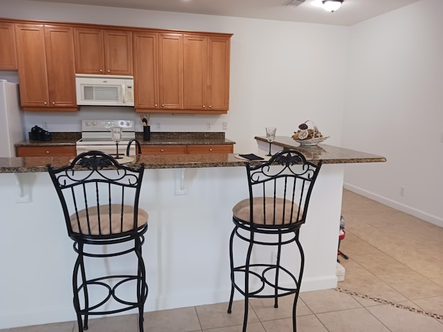 kitchen featuring a breakfast bar area, light tile patterned floors, white appliances, and dark stone counters