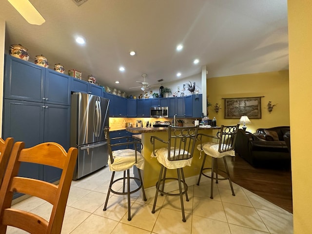 kitchen featuring light tile patterned floors, stainless steel appliances, ceiling fan, a kitchen breakfast bar, and blue cabinets