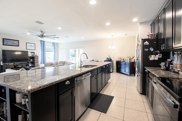 kitchen with ceiling fan with notable chandelier, stainless steel appliances, light stone counters, an island with sink, and sink