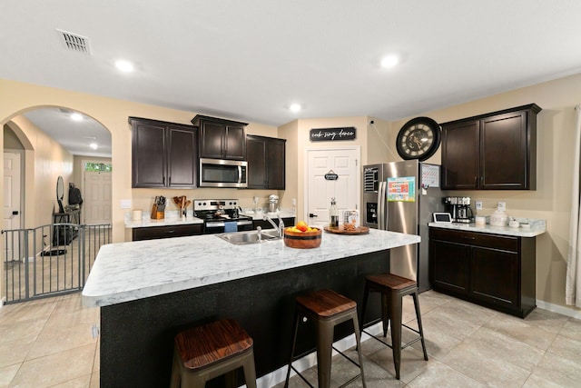 kitchen with sink, an island with sink, a breakfast bar, dark brown cabinets, and appliances with stainless steel finishes