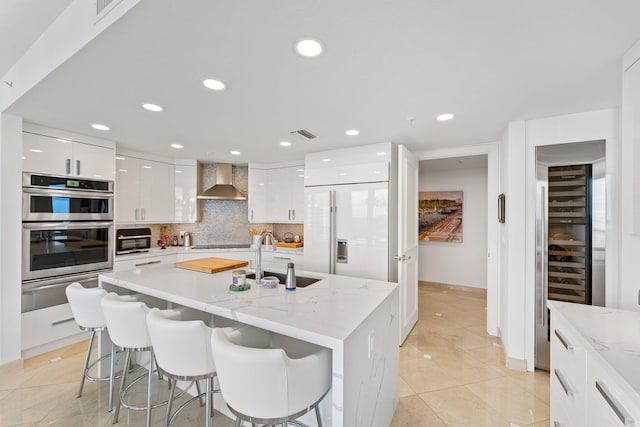 kitchen with a kitchen breakfast bar, sink, white cabinetry, wall chimney range hood, and an island with sink