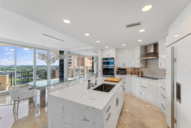 kitchen with tasteful backsplash, stainless steel double oven, wall chimney range hood, sink, and an island with sink