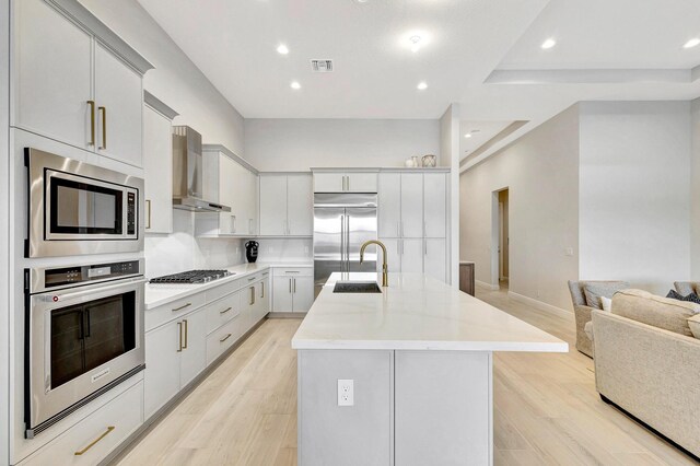 kitchen featuring built in appliances, a center island with sink, wall chimney exhaust hood, and light hardwood / wood-style floors