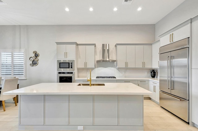 kitchen with sink, built in appliances, a kitchen island with sink, and wall chimney range hood