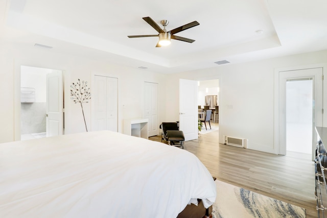 bedroom with a raised ceiling, connected bathroom, light wood-type flooring, and ceiling fan