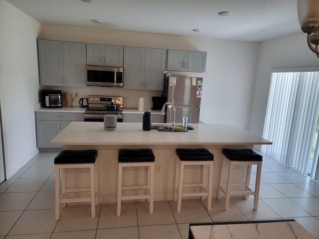 kitchen featuring gray cabinetry, light tile patterned floors, a kitchen island with sink, a breakfast bar, and stainless steel appliances