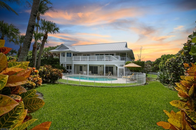 back house at dusk with a balcony, a patio, a lawn, and a fenced in pool