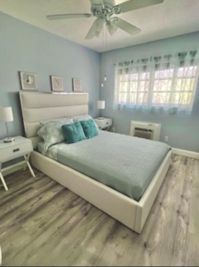 bedroom with ceiling fan, a wall unit AC, wood finished floors, and baseboards