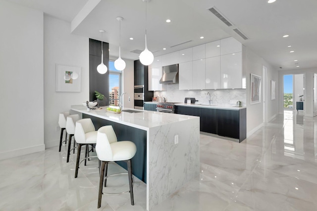 kitchen with white cabinetry, backsplash, a breakfast bar area, decorative light fixtures, and wall chimney range hood