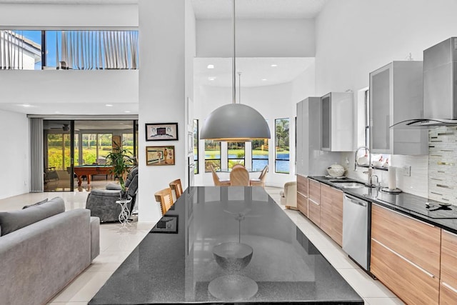 kitchen featuring dark stone counters, dishwasher, sink, wall chimney exhaust hood, and a towering ceiling