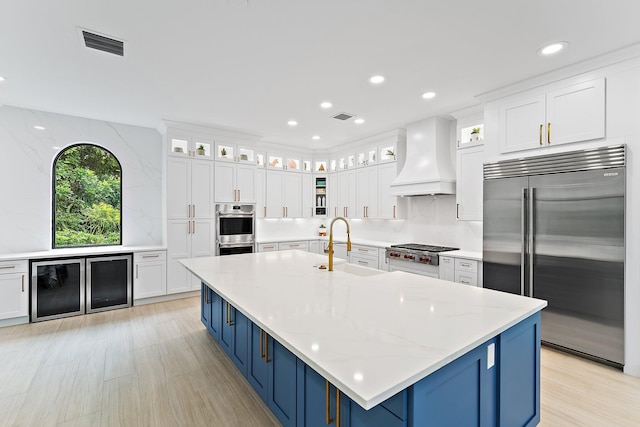 kitchen with glass insert cabinets, custom exhaust hood, appliances with stainless steel finishes, and blue cabinetry