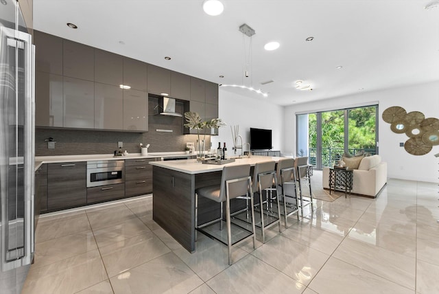 kitchen with decorative backsplash, an island with sink, wall chimney exhaust hood, dark brown cabinetry, and stainless steel appliances