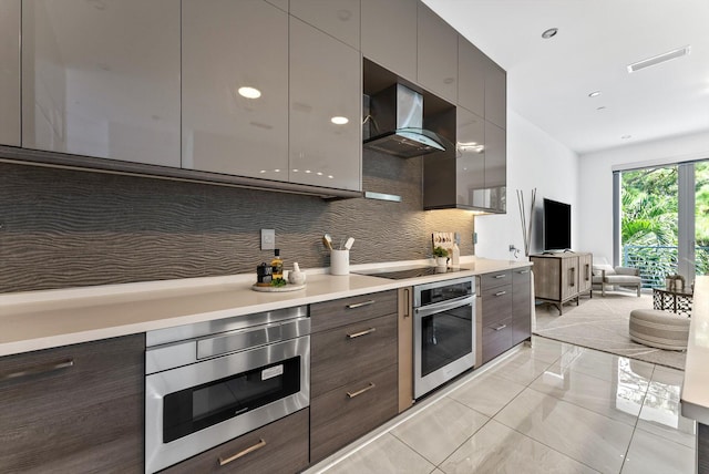 kitchen featuring stainless steel appliances, wall chimney range hood, dark brown cabinetry, and tasteful backsplash