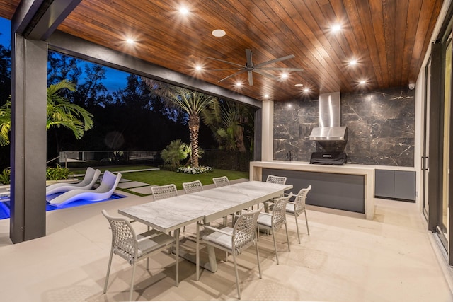 patio at night with ceiling fan, a swimming pool, a lawn, and exterior kitchen