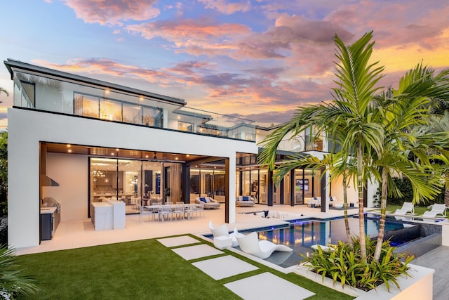 back house at dusk featuring a lawn, a balcony, an outdoor kitchen, and a patio area