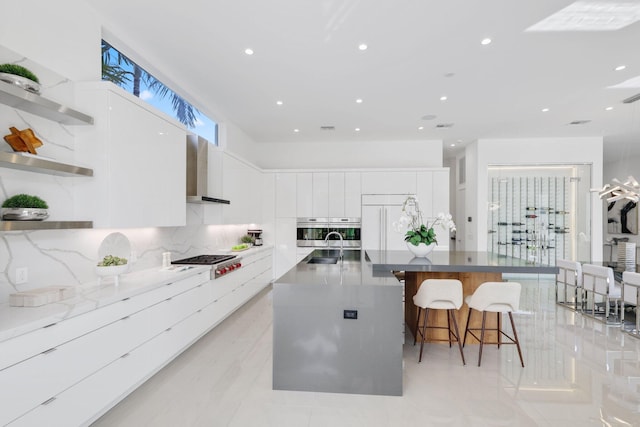 kitchen featuring wall chimney exhaust hood, a large island with sink, white cabinetry, stainless steel appliances, and a kitchen bar