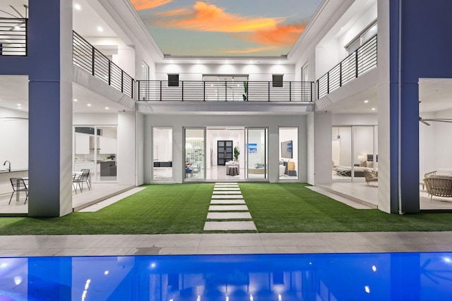 back house at dusk with a patio, a balcony, and a lawn