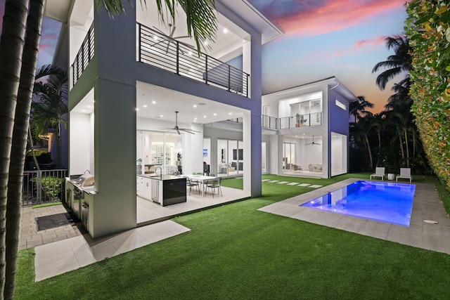 back house at dusk featuring a yard, a jacuzzi, a balcony, exterior kitchen, and a patio area