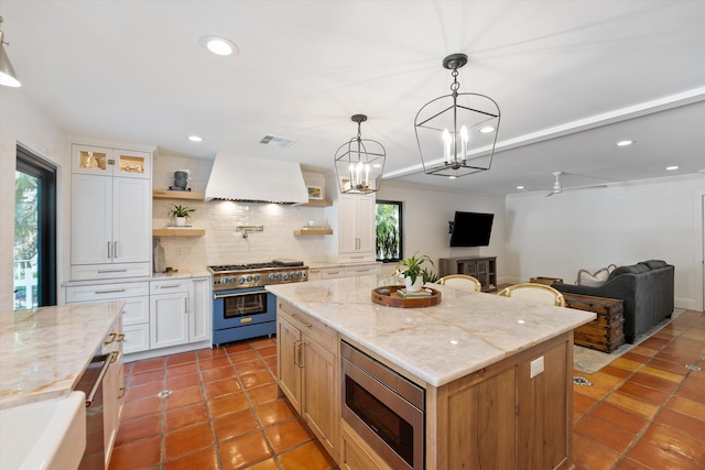 kitchen with a kitchen island, white cabinetry, appliances with stainless steel finishes, custom range hood, and ceiling fan with notable chandelier