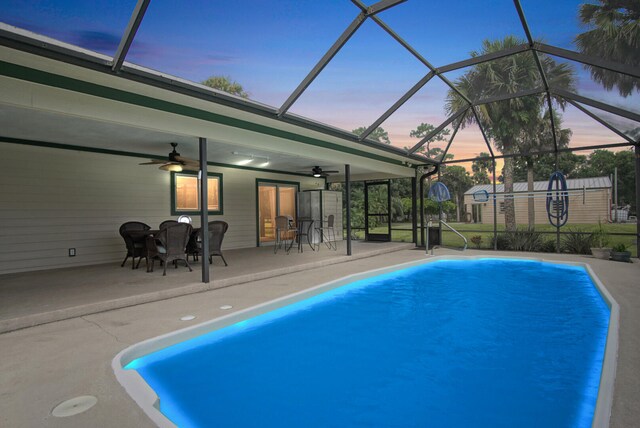 pool at dusk with glass enclosure, ceiling fan, a shed, and a patio area