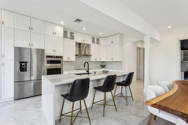 kitchen with white cabinets, a center island with sink, stainless steel appliances, and wall chimney range hood