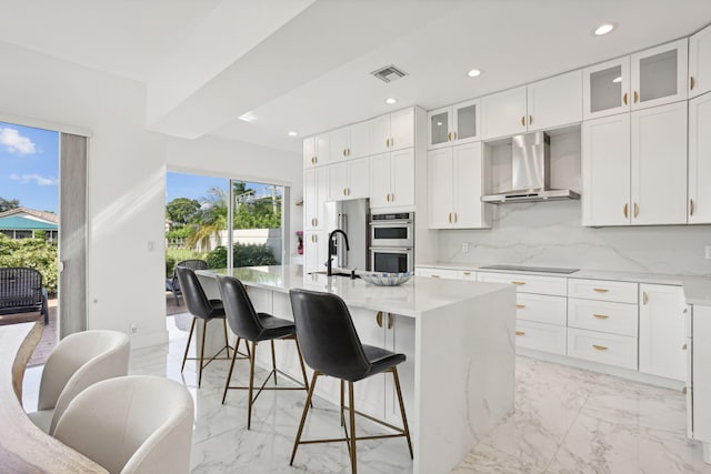 kitchen featuring white cabinets, an island with sink, wall chimney range hood, appliances with stainless steel finishes, and decorative backsplash