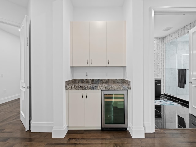 bar with dark wood-type flooring, wine cooler, white cabinetry, and dark stone counters