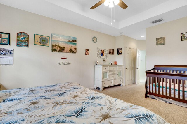 bedroom with a raised ceiling, ceiling fan, light colored carpet, and a nursery area