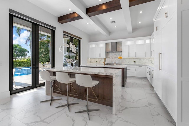 kitchen featuring white cabinetry, backsplash, wall chimney range hood, beamed ceiling, and a kitchen island with sink