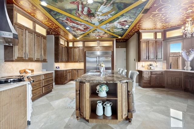 kitchen featuring light stone countertops, backsplash, wall chimney range hood, a center island with sink, and stainless steel built in fridge