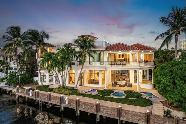 back house at dusk with an outdoor hangout area, a balcony, a pool with hot tub, a water view, and a patio