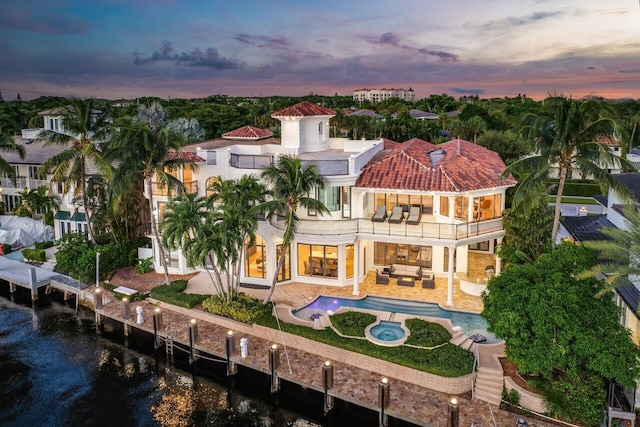 back house at dusk featuring an outdoor living space, a water view, a swimming pool with hot tub, a balcony, and a patio