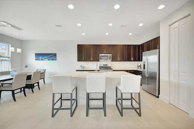 kitchen featuring light tile patterned flooring, hanging light fixtures, a kitchen island with sink, stainless steel appliances, and a breakfast bar area