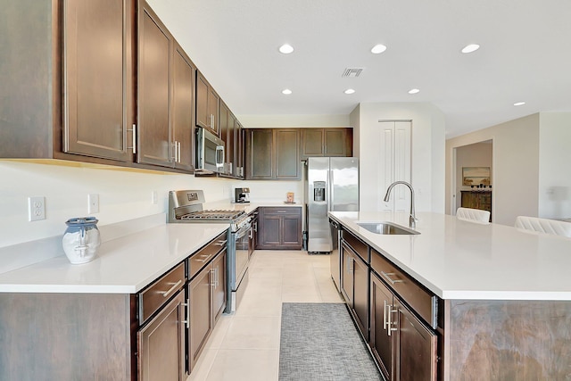 kitchen featuring appliances with stainless steel finishes, an island with sink, light tile patterned floors, dark brown cabinetry, and sink