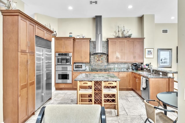 kitchen featuring wall chimney exhaust hood, decorative backsplash, appliances with stainless steel finishes, a kitchen island, and light stone counters